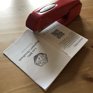 The image shows a small DIY booklet titled *Law RPG Beginner’s Handbook* being stapled along the fold with a red Swingline stapler. The booklet is opened to display the back cover and inside back page, which includes a QR code labeled “Interactive Helper” and instructions for accessing online resources. The front cover features a 20-sided die illustration and the author’s name, David Colarusso. The booklet rests on a wooden surface, and the lighting is soft, creating a warm, casual crafting atmosphere. This moment captures the final step of assembling a printed zine or mini-handbook, emphasizing its homemade, accessible nature.